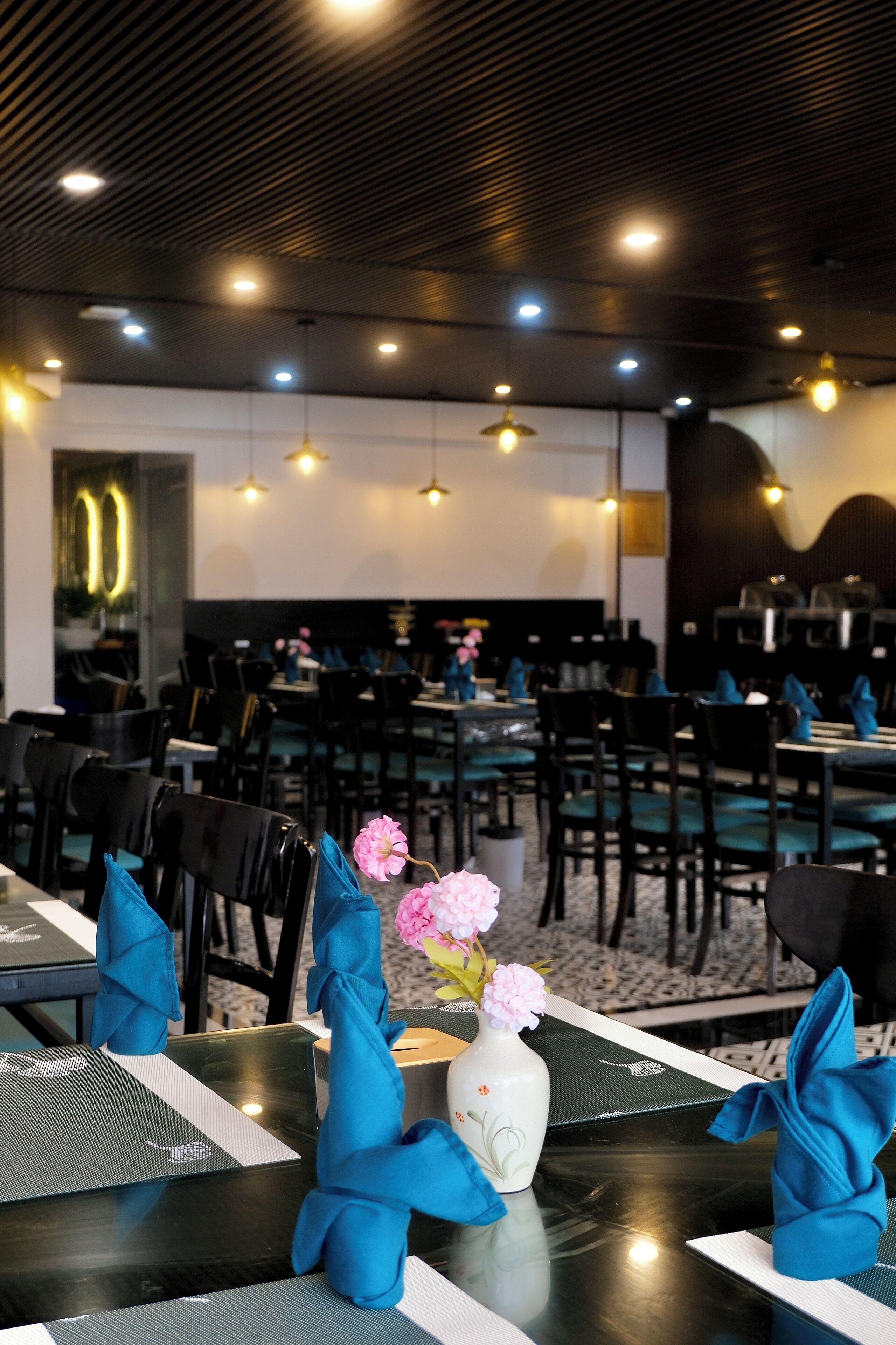 A restaurant with tables set with blue napkins, a vase with pink flowers, and a modern ceiling with recessed lighting.