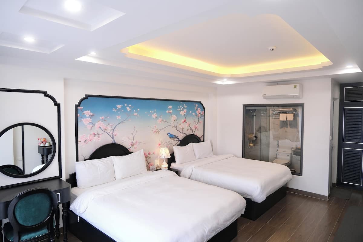 A hotel room with two beds, a desk with a chair, a large mirror, a wall-mounted air conditioner, and a bathroom with a glass door.
