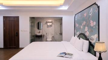 Habitación de hotel con una cama grande, una mesita de noche con lámpara, un cuadro de pájaros en la pared, una puerta y un baño visible a través de una puerta de cristal.