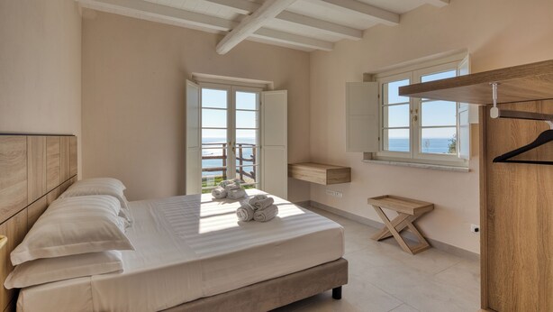 Een slaapkamer met een groot bed, houten hoofdbord, een houten nachtkastje, een klein houten krukje, een houten plank en een raam met uitzicht op zee.