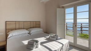 Een slaapkamer met een groot bed, een houten hoofdbord, uitzicht op zee door een glazen deur en een bankje bij de deur.
