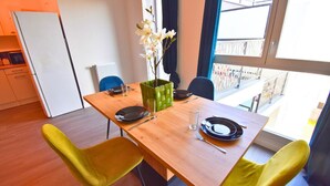 Un coin repas avec une table en bois, deux chaises jaunes, un vase vert contenant des fleurs blanches et, par la fenêtre, une vue sur un balcon.