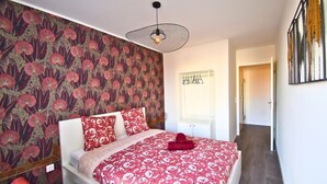 Une chambre avec un papier peint à motifs, un lit avec une literie rouge, un luminaire suspendu et une porte donnant sur une autre pièce.