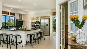 Uma cozinha moderna com uma ilha central, banquetas, eletrodomésticos de aço inoxidável e grandes janelas.