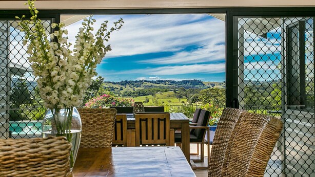 Uma área de refeições com mesa e cadeiras, um vaso com flores e uma vista panorâmica por grandes janelas.