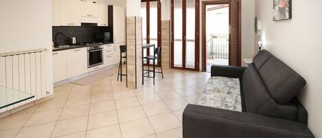 A modern kitchen with white cabinets, a black countertop, and a built-in oven. There is a dining area with a black table and two chairs. A black leather sofa is in the living area, and a glass table is in the foreground.