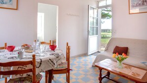 A dining area with a table set for two, a sofa, and a window with a view of greenery. - Rose-May by Interhome (La Bernerie-en Retz)