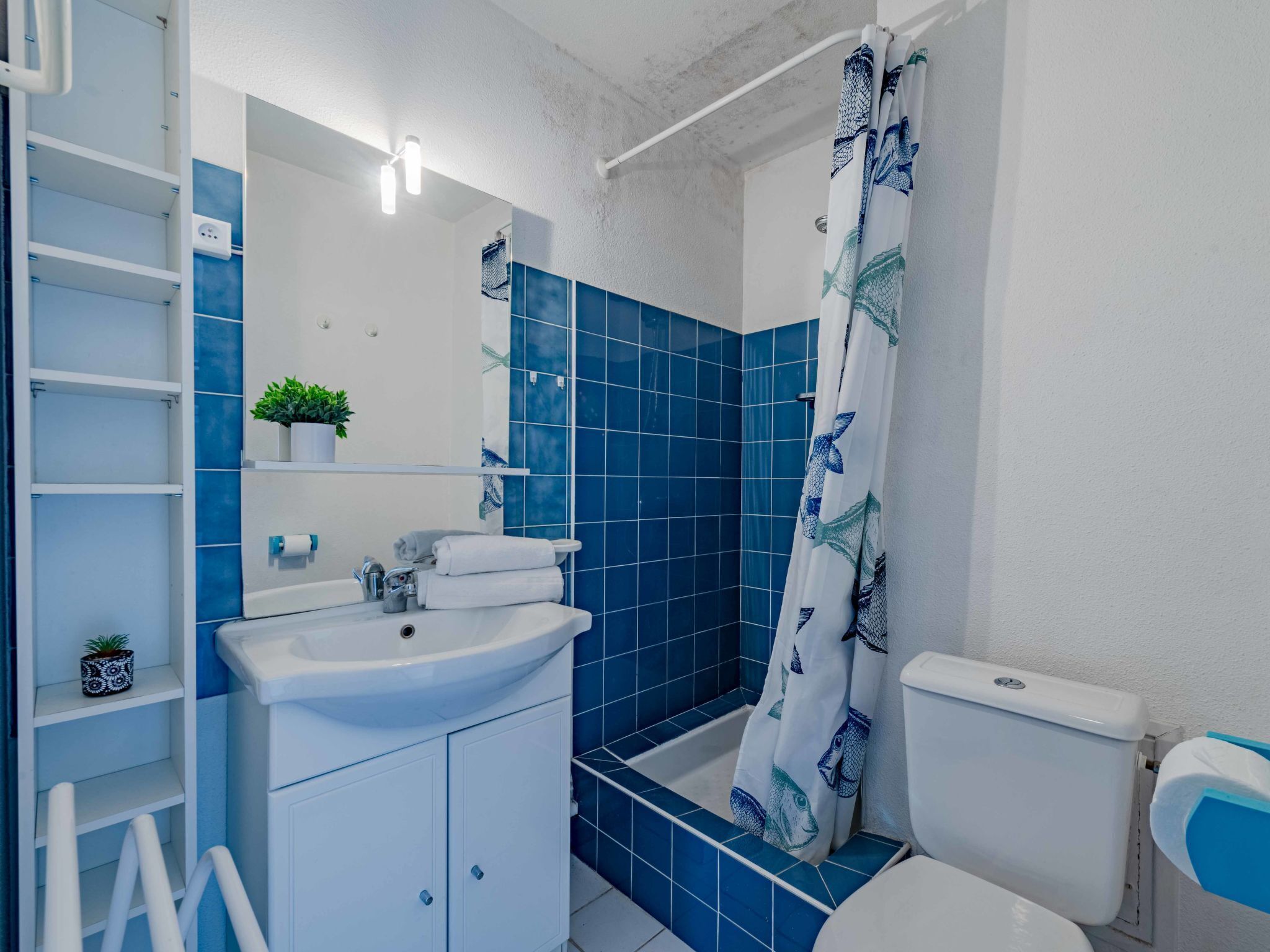 Um banheiro com pia branca, azulejos azuis, cortina de chuveiro com desenhos de peixes e vaso sanitário.