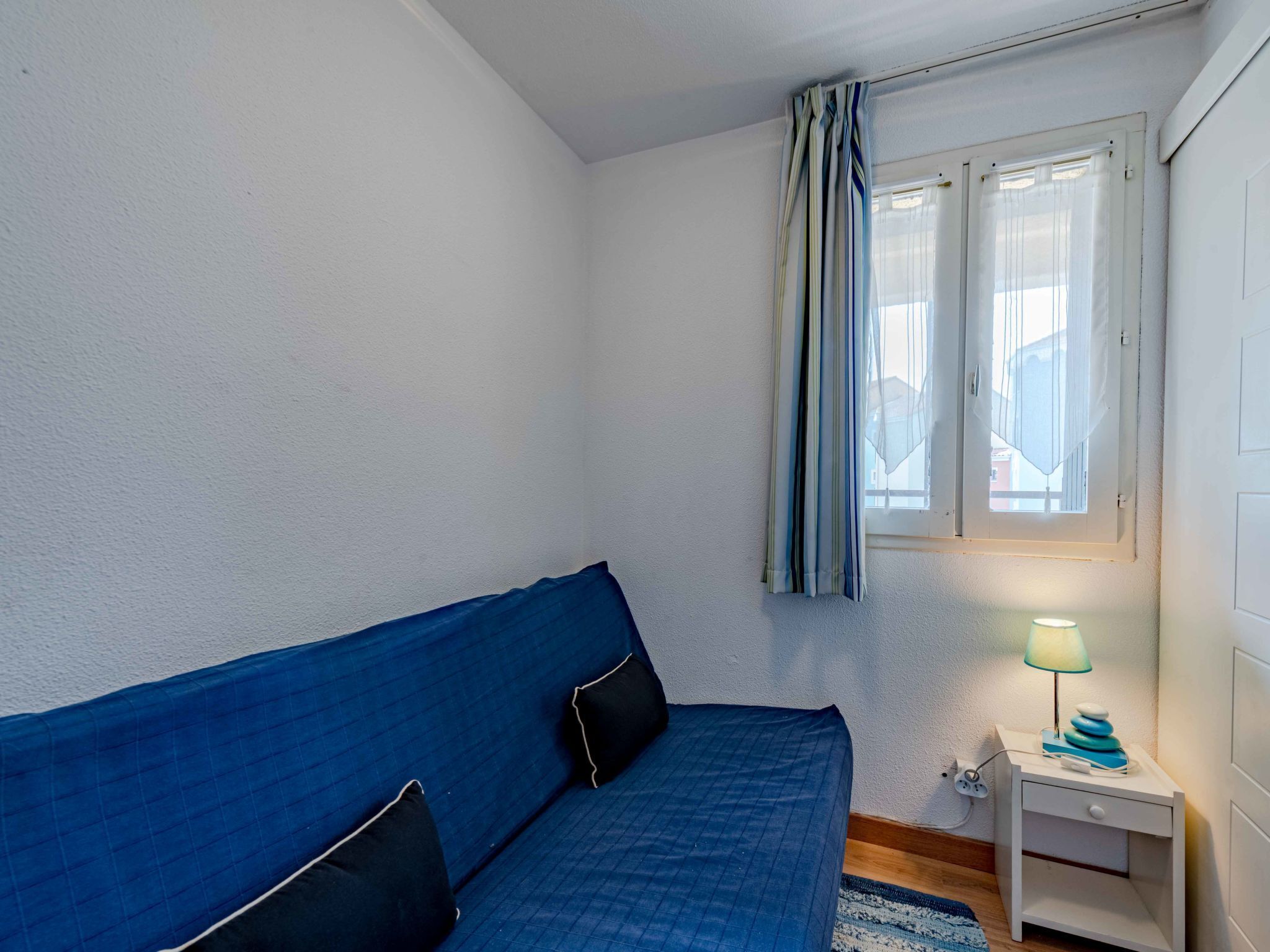 A bedroom with a blue bed, a nightstand with a lamp, and a window with curtains.