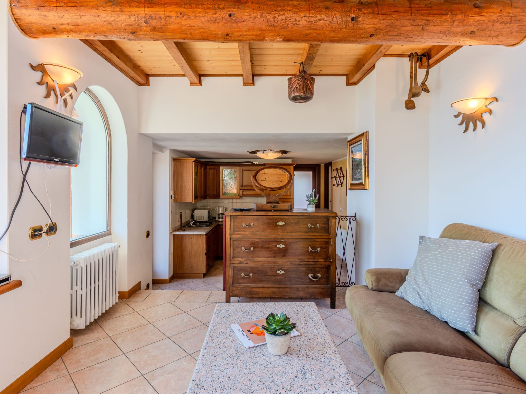 Een gezellige woonkamer met een houten plafond, een bank, een bijzettafeltje met een plant en een keuken op de achtergrond.