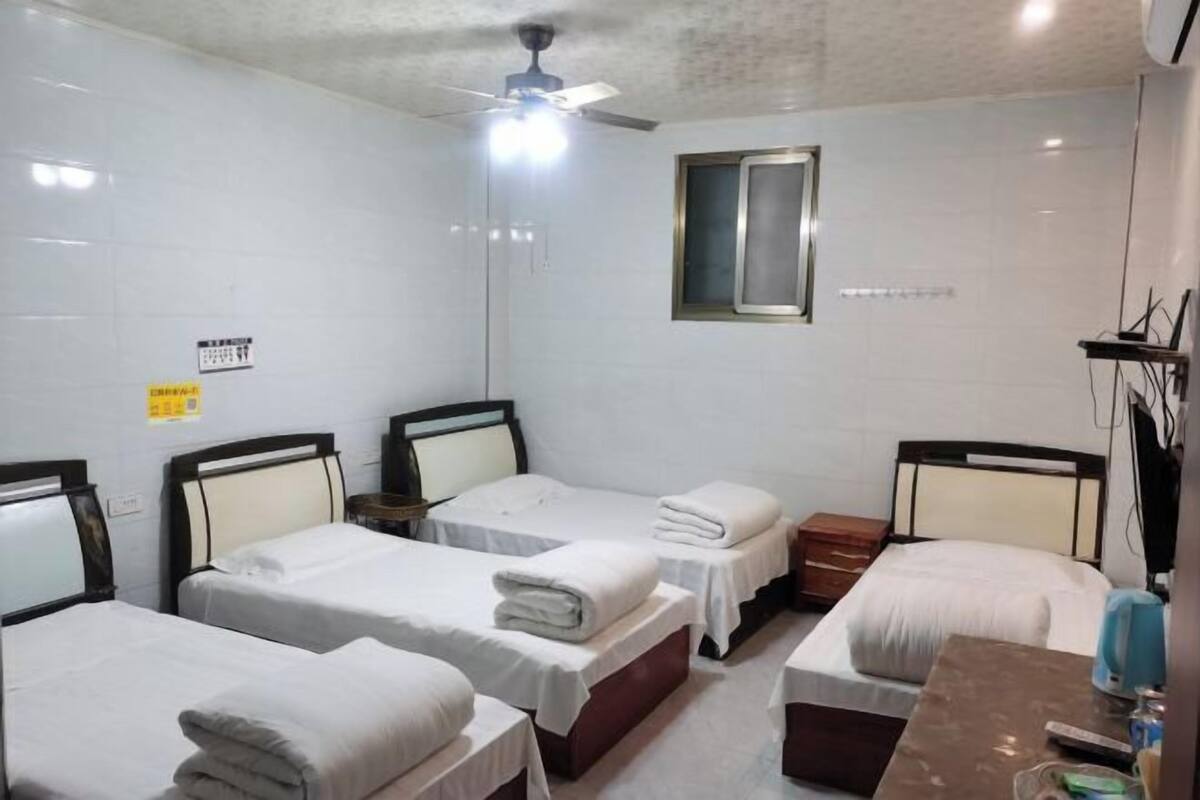 A hotel room with four single beds, a ceiling fan, a window, and a small table with a lamp.