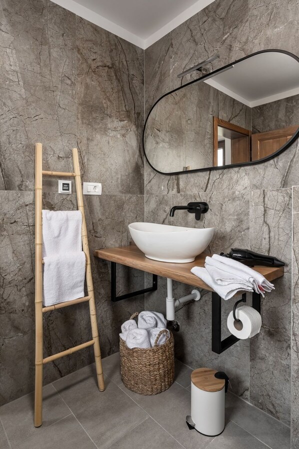 A bathroom with a marble wall, a wooden vanity, a white sink, a bamboo ladder, a basket, and a toilet paper holder.