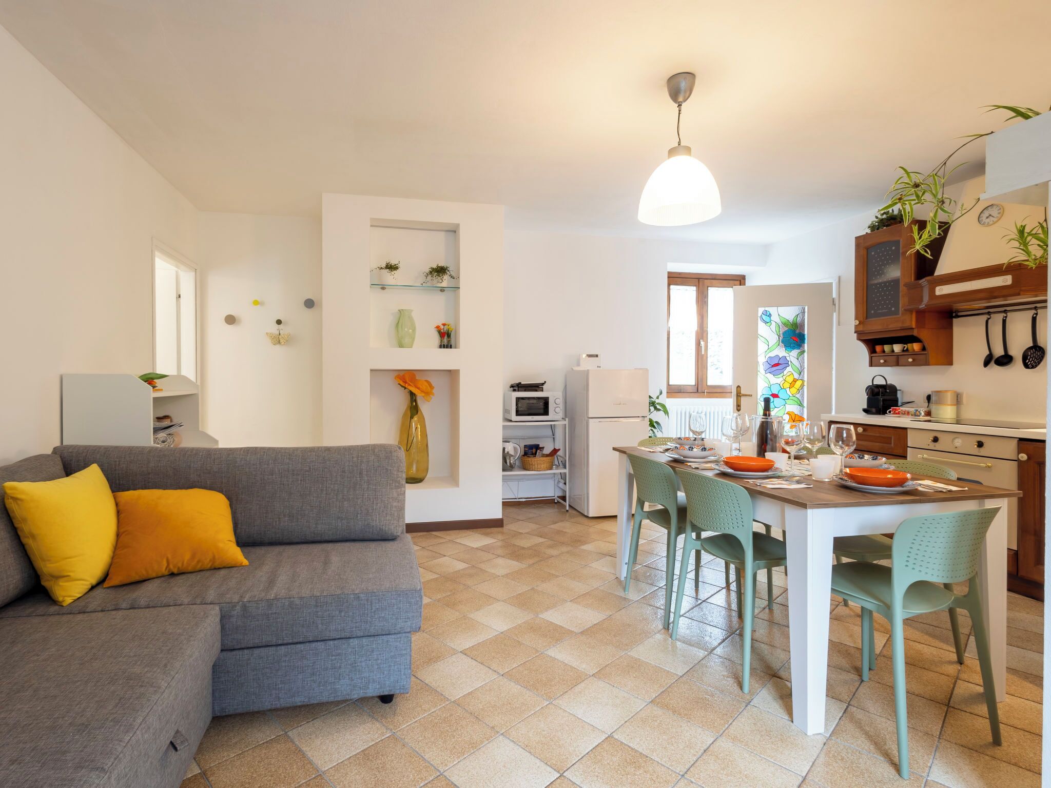 Een moderne woonkamer met een grijze bank, een eettafel met stoelen, een keuken met een koelkast, magnetron en kastjes, en een hanglamp.