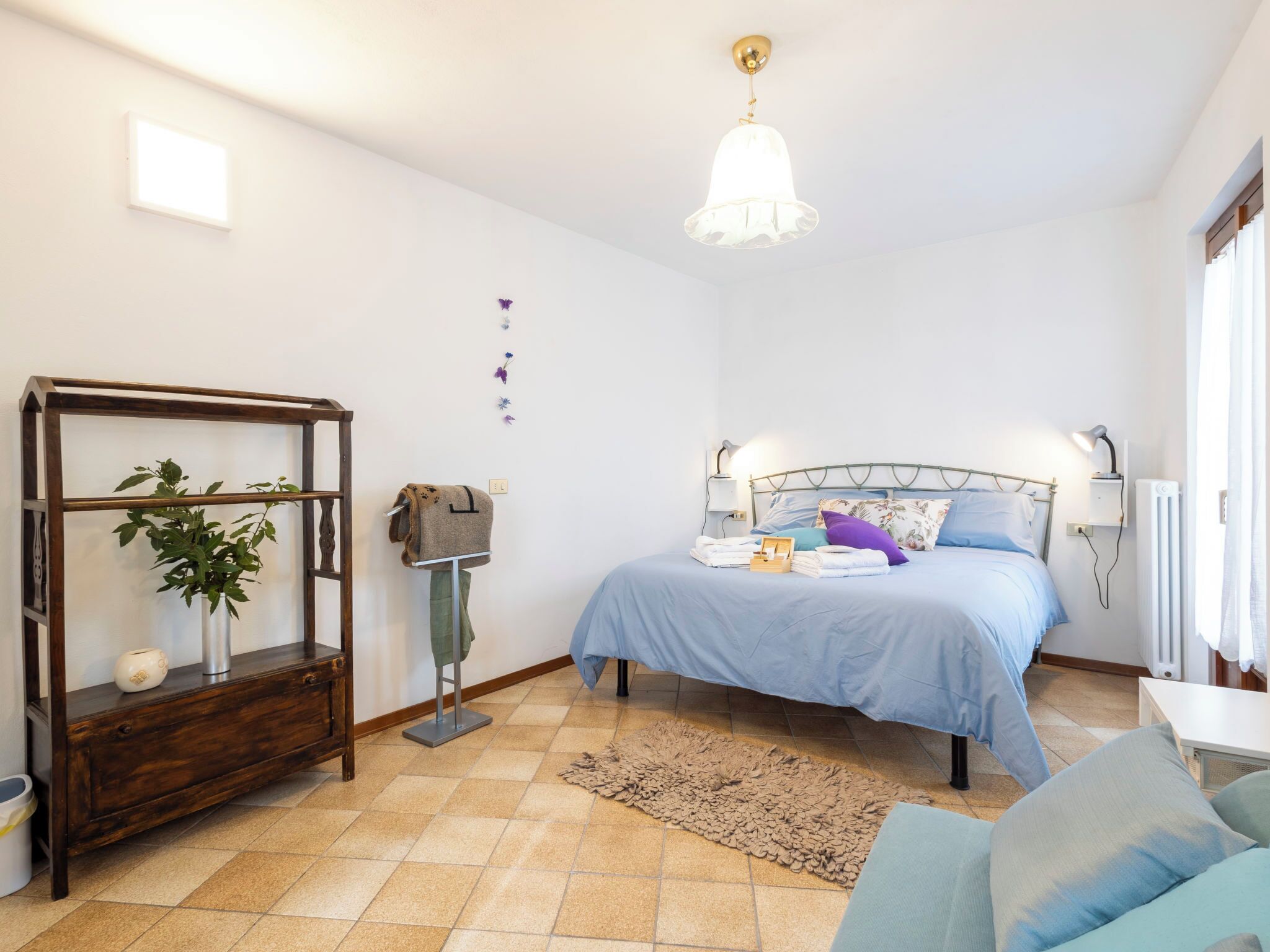 A bedroom with a bed, bedside lamps, a hanging light fixture, a wooden dresser, a plant, and a radiator.