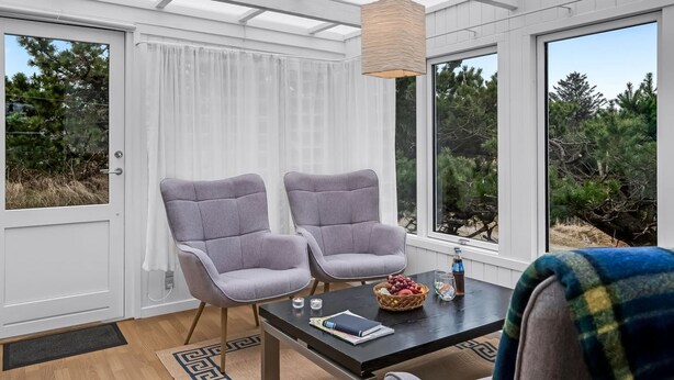A conservatory with two grey armchairs, a dark wooden table with a book, glasses, and a basket of fruit.
