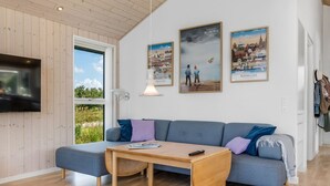 Ein modernes Wohnzimmer mit Holzboden, einem grauen Sofa, einem Couchtisch aus Holz und einem an der Wand montierten Fernseher. An der Wand hängen drei gerahmte Bilder, und ein Fenster bietet Blick auf Grünflächen.