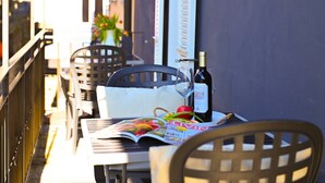 En balkon med et bord dækket til to, med en vinflaske, et glas, et magasin og en kurv med frugt.