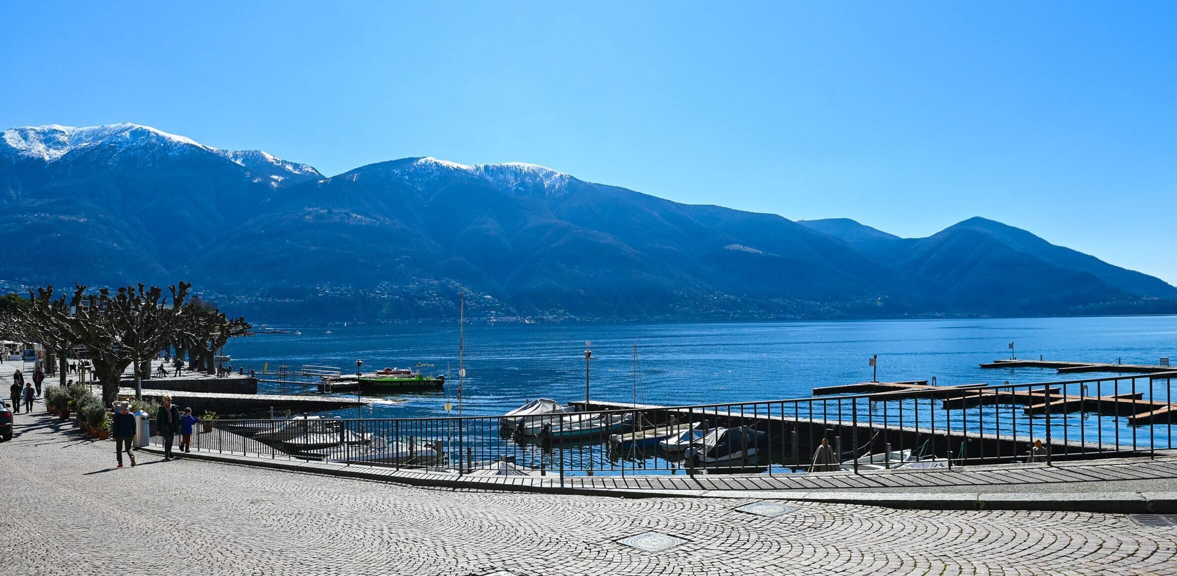 Une promenade en bord de mer avec un chemin pavé, des bateaux amarrés au quai et une chaîne de montagnes en arrière-plan.