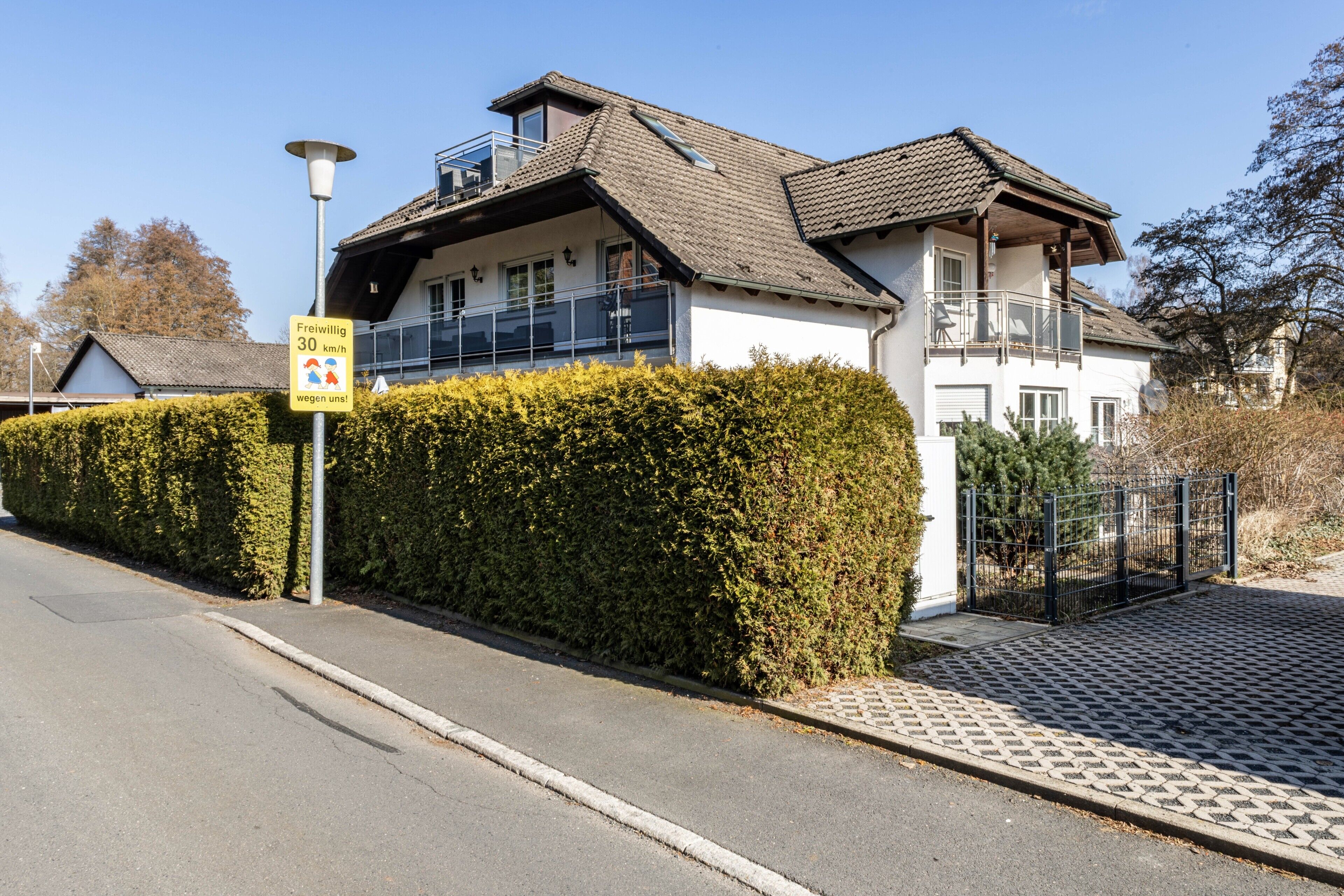 A two-story house with a balcony, a paved driveway, and a hedge along the street.