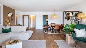 Ein modernes Wohnzimmer mit Sofa, Esstisch und Stühlen, einem Bücherregal mit Pflanzen und einem großen Spiegel.