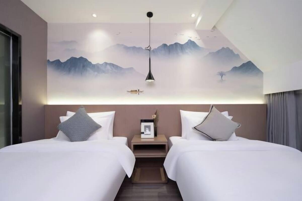 A hotel room with two beds, a nightstand, a framed picture, and a wall mural of mountains.