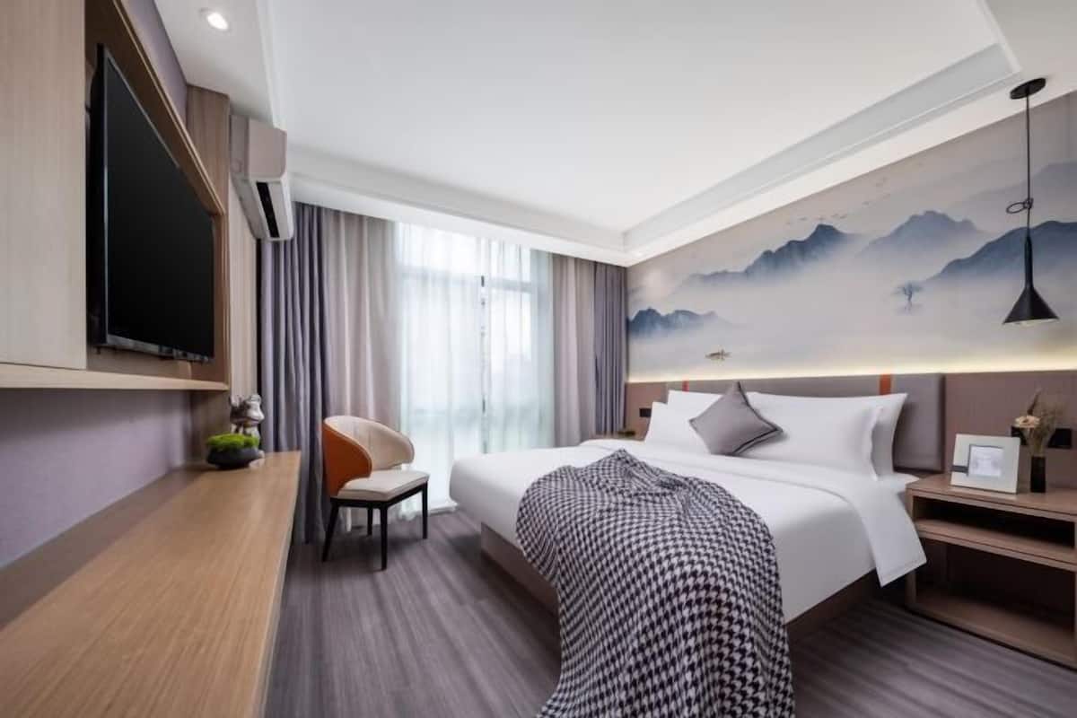 A hotel room with a bed, a chair, a TV, and a wall with a mountain painting.