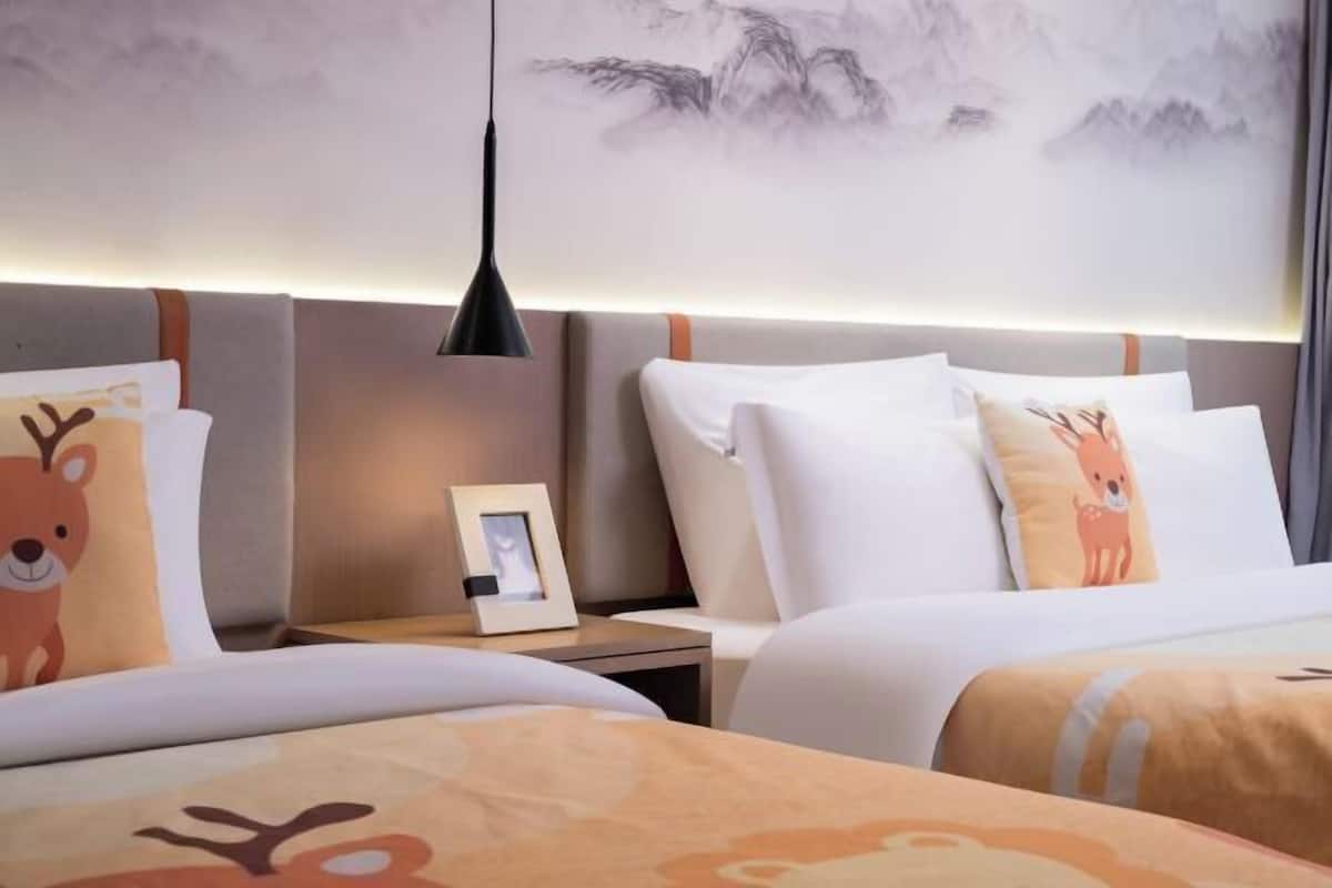 A hotel room with two beds, a nightstand with a picture frame, and a wall with a mountain painting.