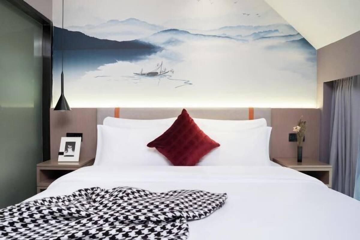 A hotel room with a large bed, a red pillow, a black and white patterned blanket, a nightstand with a framed photo, and a wall painting of a serene landscape.