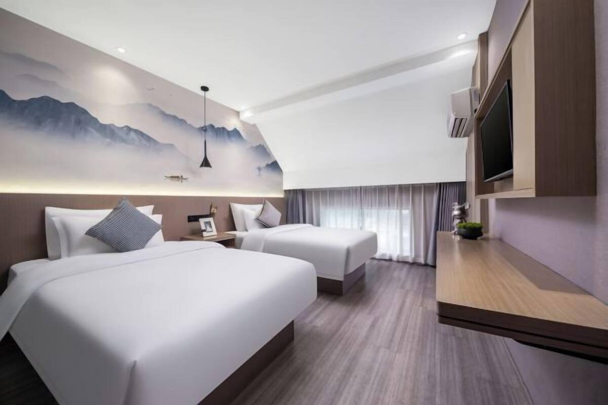 Une chambre d’hôtel avec deux lits, une télévision, un climatiseur fixé au mur et un tableau représentant des montagnes.