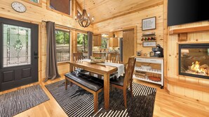 Acogedor interior de una cabaña de madera con comedor, chimenea y cocina visible a través de una puerta abierta.