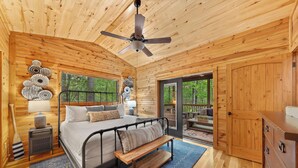 Dormitorio de una cabaña de madera con cama, banca, ventilador de techo y vista al exterior.