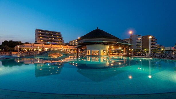 A swimming pool with a thatched-roof structure in the center, surrounded by lounge chairs and a building with multiple floors and balconies. - Chic double room near the Bibione coast (Bibione)