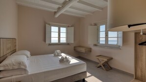 Une chambre à coucher comprenant un lit, deux fenêtres, une tête de lit en bois, une petite table en bois et un plafond avec des poutres apparentes.