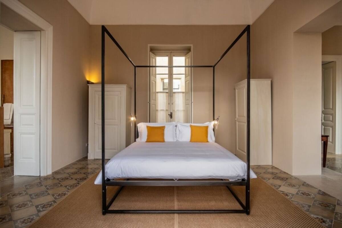Un lit à baldaquin avec du linge de lit blanc et des coussins orange, un tapis tissé et une vue sur un couloir avec une porte et une fenêtre.