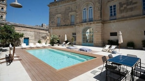 A pool area with lounge chairs and a table, surrounded by a historic building with a fountain and a bell tower. - PALAZZO GARZIA - Dimora Storica (Maglie)
