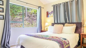 A bedroom with a large bed, a window with curtains, a nightstand, and a painting on the wall. - Annabells Bed and Breakfast (Whitianga)