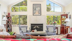 A spacious living room with a fireplace, two armchairs, a sofa, and a large window overlooking a garden. - Annabells Bed and Breakfast (Whitianga)