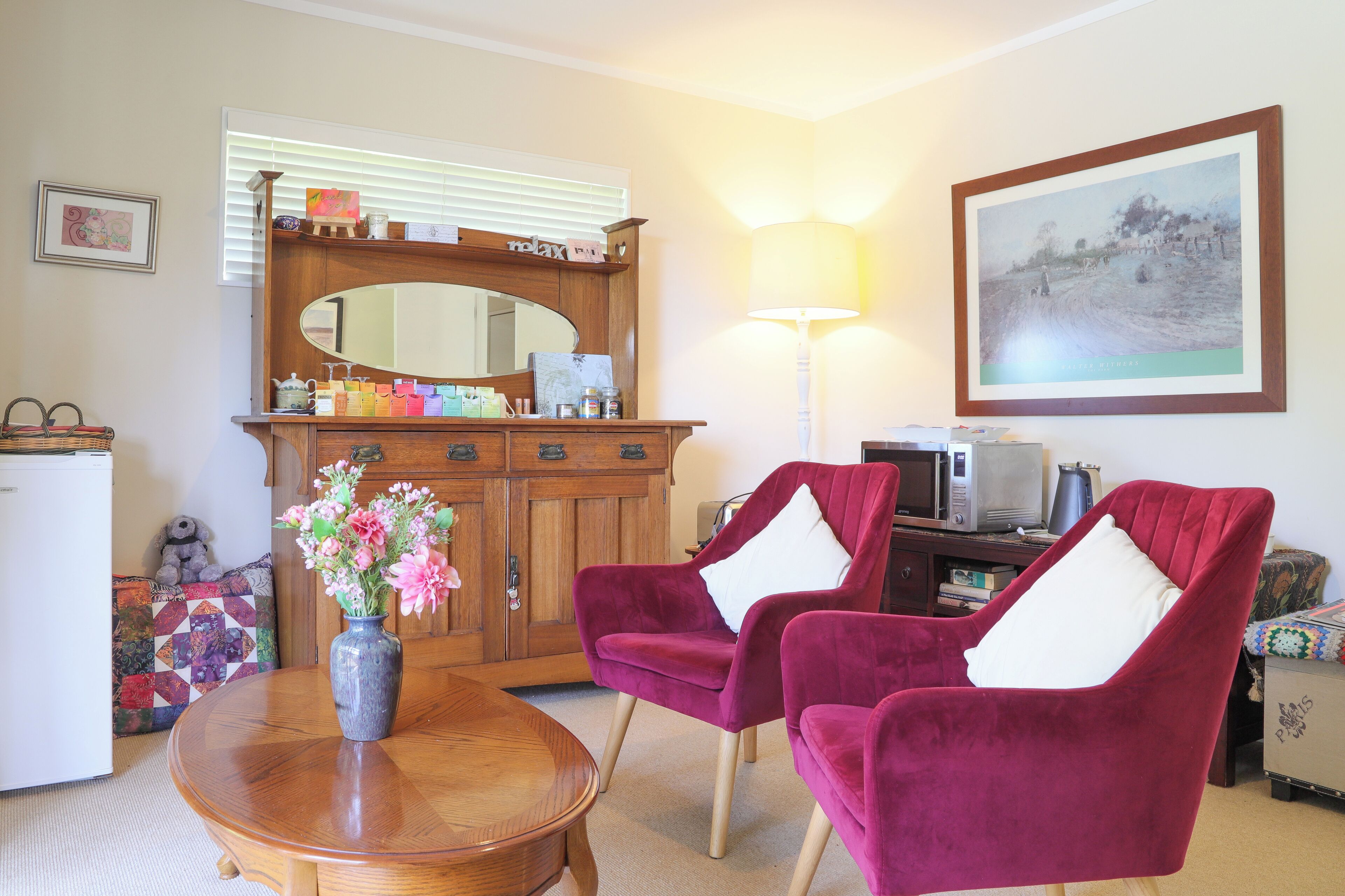 A living room with a wooden cabinet, two purple armchairs, a round wooden table, a vase with flowers, and a framed landscape painting on the wall.