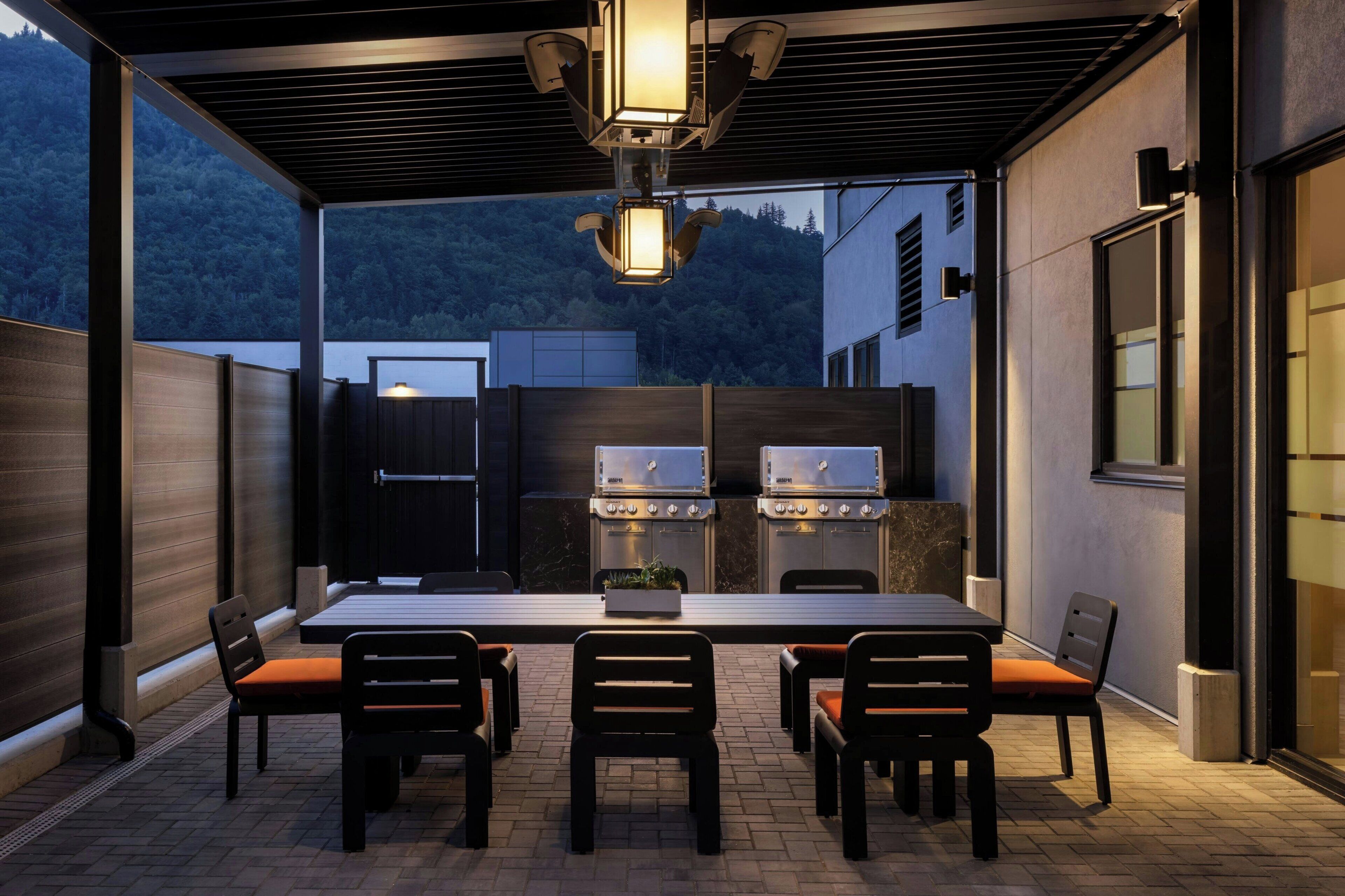An outdoor patio with a dining table and chairs, a grill, and a view of a forested hillside.
