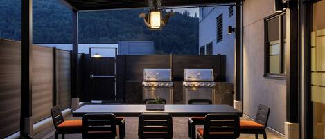 An outdoor patio with a dining table and chairs, a grill, and a view of a forested hillside.