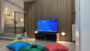 Moderni olohuone, jossa on litteänäyttöinen televisio, jolla näkyy videopeli, geometrisesti muotoiltu kaide portaikossa ja värikkäitä koristetyynyjä lattialla.