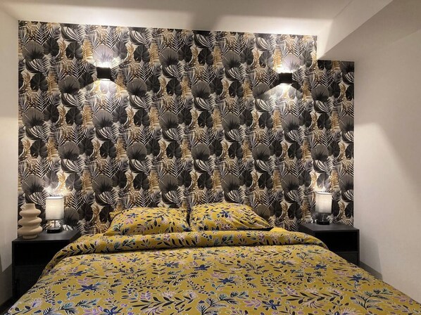 A bedroom with a patterned wallpaper, a bed with a floral bedspread, two bedside lamps, and a nightstand with a vase.