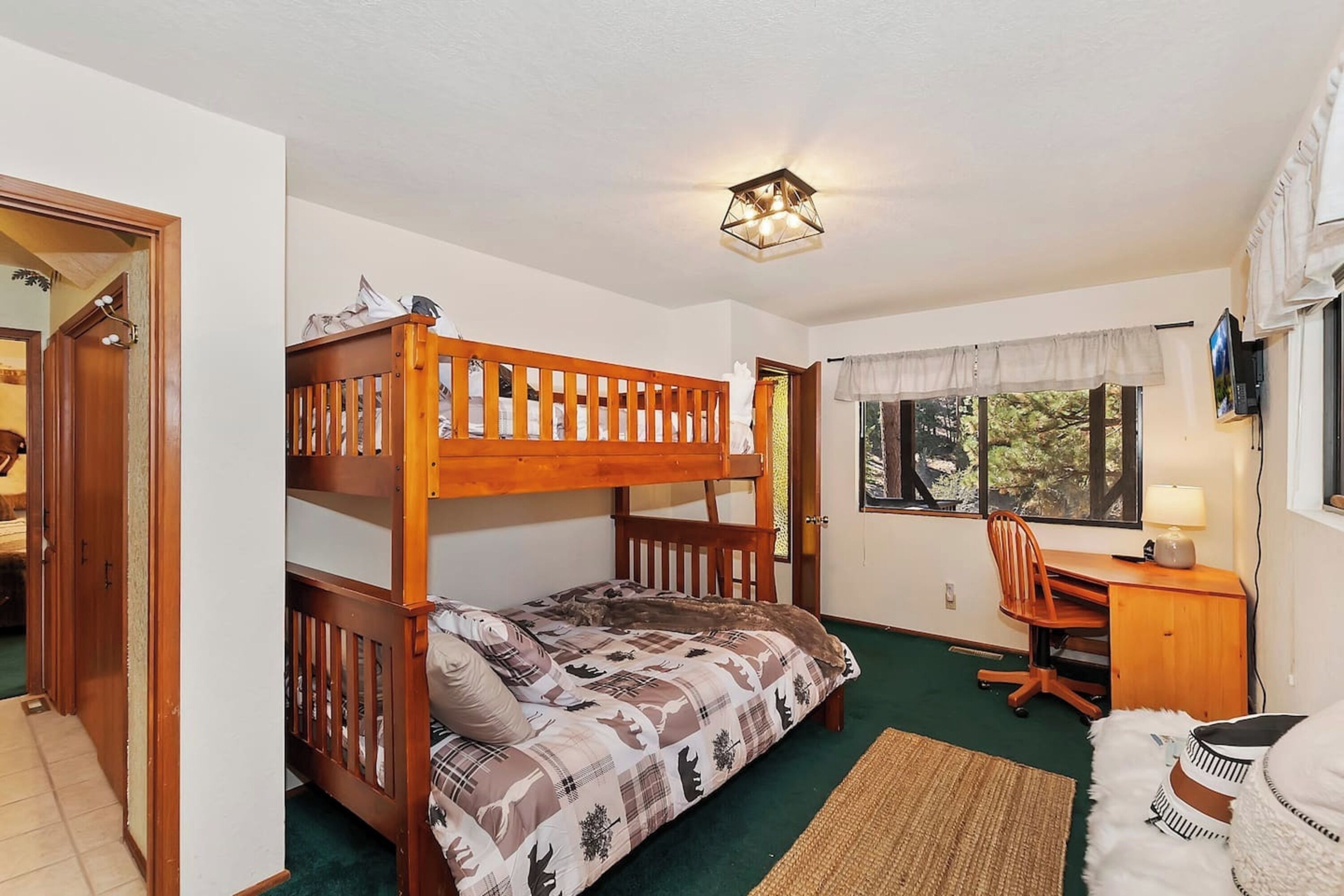 A bunk bed room with a desk and chair, a window with a view, and a door leading to another room.