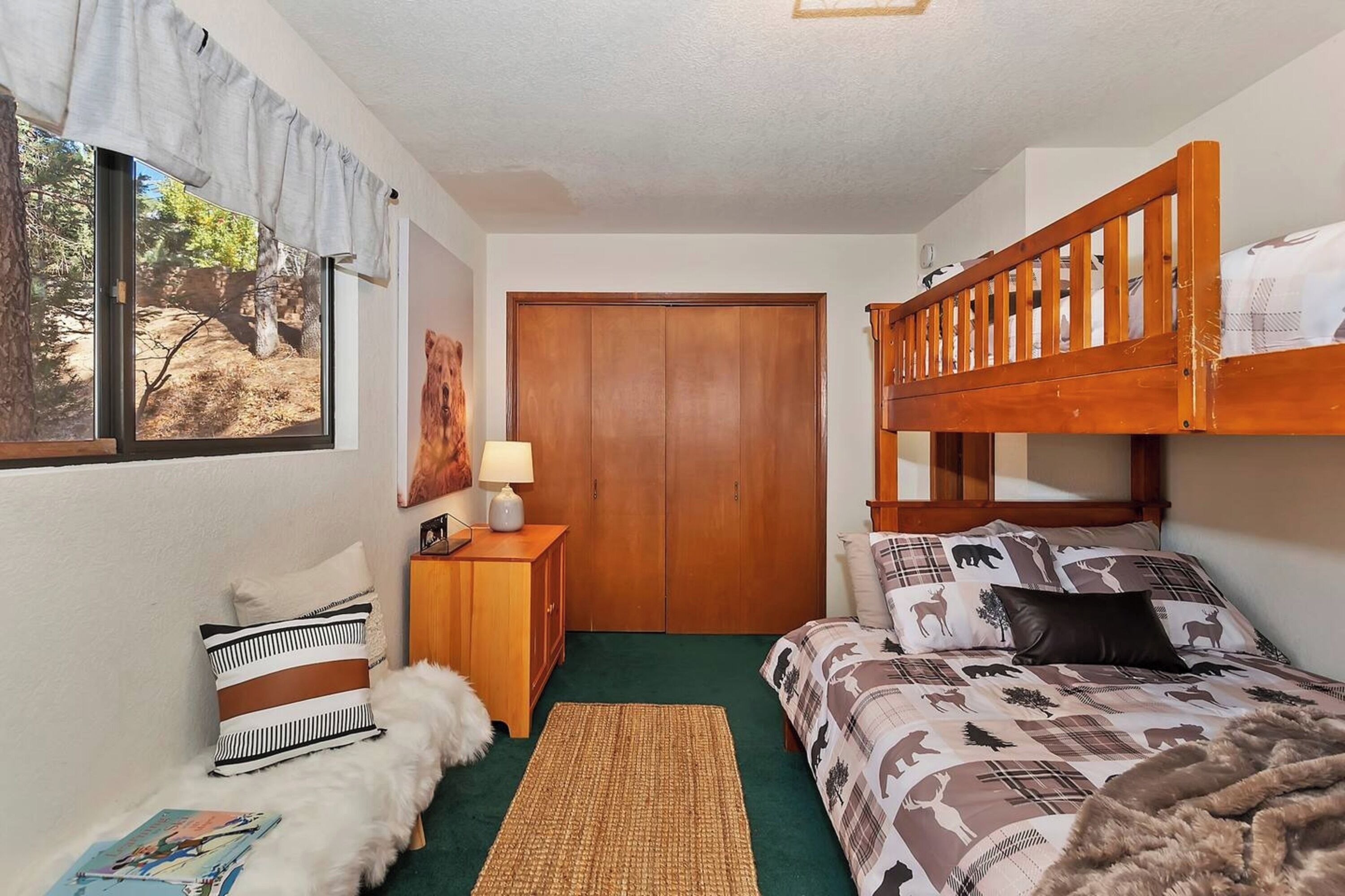 A bedroom with a bunk bed, a wooden wardrobe, a window with a view of trees, a lamp, and a rug.