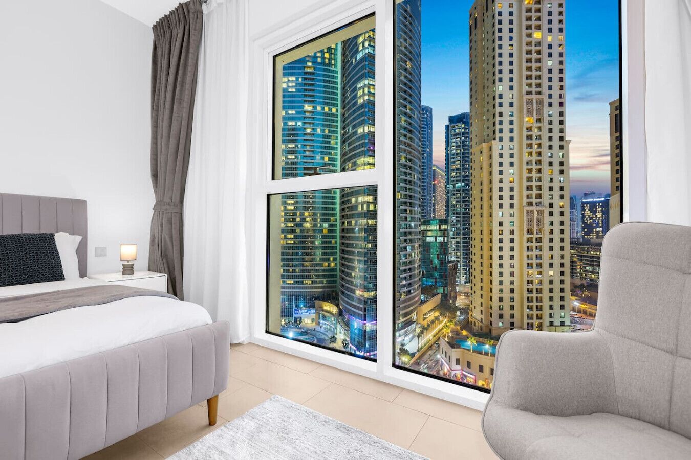 Camera d'hotel moderna con un'ampia finestra che si affaccia sulla città, un letto, un comodino con una lampada e una poltrona.