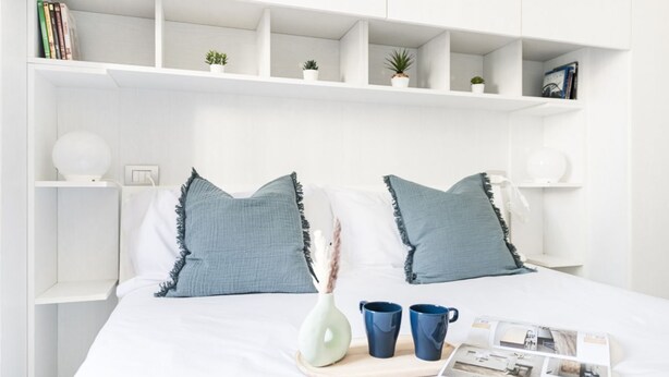 A neatly arranged bedroom with a bed, bedside tables, shelves with books and plants, and a tray with a vase and mugs.