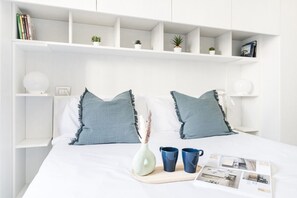 Une chambre à coucher bien rangée, avec un lit, des tables de chevet, des étagères remplies de livres et de plantes, et un plateau sur lequel se trouvent un vase et des tasses.