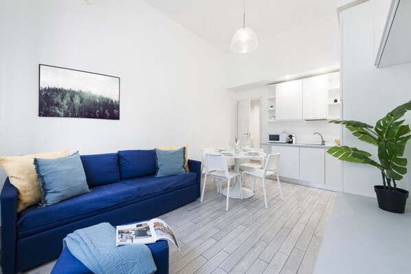 A modern living room with a blue sofa, a dining table with chairs, a kitchen area, and a large potted plant. - Easylife - Elegant Apartment in Città Studi (Milano)