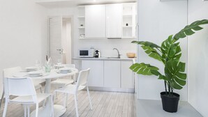 A modern kitchen with white cabinetry, a microwave, and a sink. There is a dining table with white chairs and a potted plant.