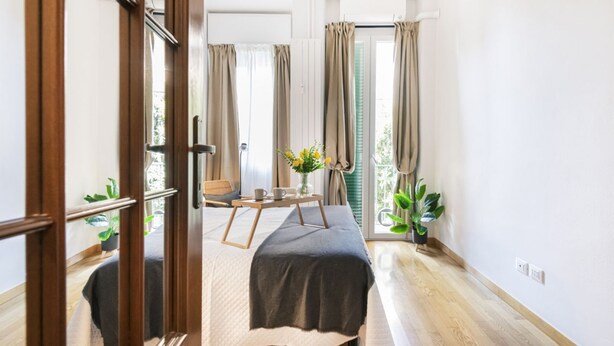 Un dormitorio con cama, una mesita de noche con un florero y una ventana con cortinas.